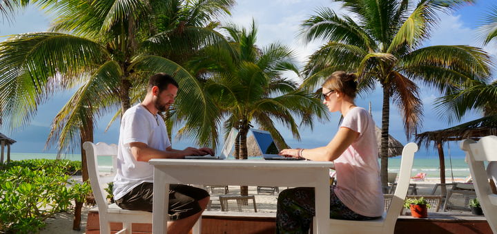 Nick and Dariece of Goats On The Road working from their laptops in front of a beautiful beach with palm trees in the background.