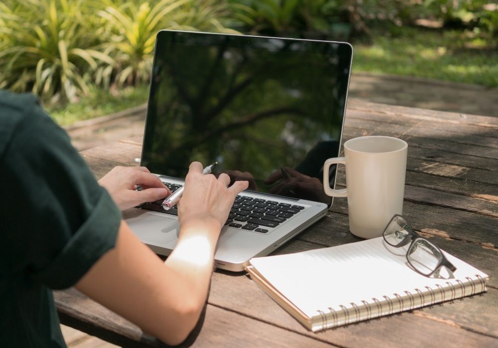 woman writing on a laptop outside at a desk with a notepad, her glasses and a coffee mug. writing and getting paid to travel