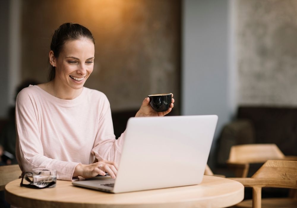 A woman working happily from her laptop in her home with an espresso in her hand