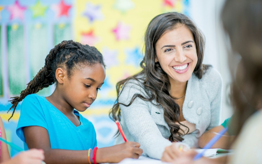 A group of elementary students are indoors in their classroom while taking notes in front of their teacher.