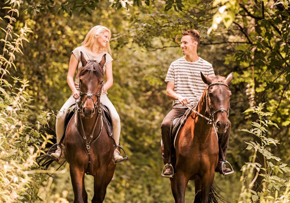 A happy couple is looking at each other while horseback riding in the countryside. 