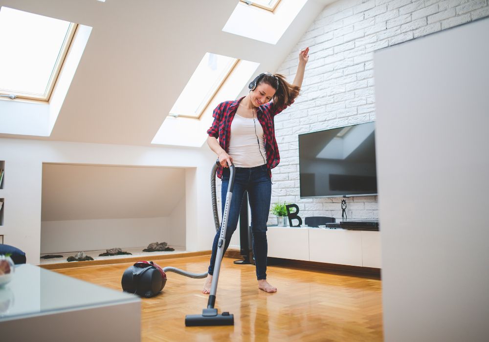 The young woman house sitter is using a vacuum cleaner to clean the house while listening to music.