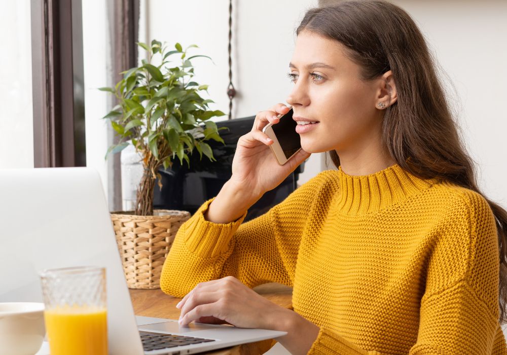 A young female entrepreneur is multitasking, talking on her mobile phone and working on her laptop as a digital nomad.