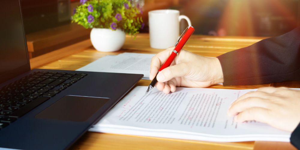 Example of person proofreading on paper with a laptop in front of them on a desktop with coffee