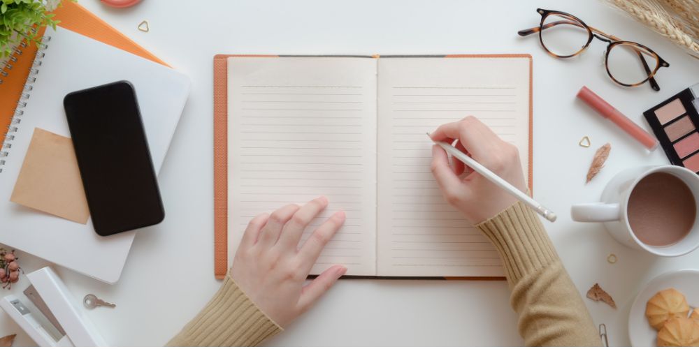 Writing in a notebook on a desk containing glasses, coffee, a phone, and more