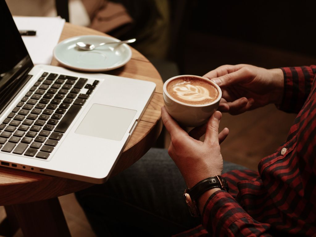 person sitting at a table with a laptop and a cup of coffee in their hand