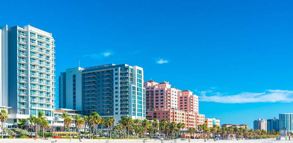 A sunny day with buildings on the background and coconut trees near the white sand
