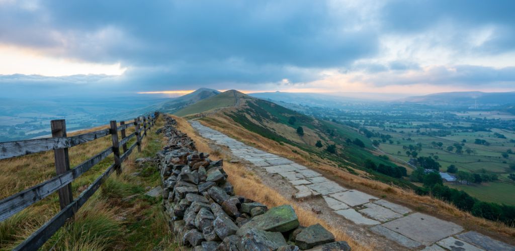 Mam Tor (Peak District National Park) with cloudy dark afternoon sky and a yellow sky caused by sunset rays with fences on the side and stones beside a tiled trekking way
 