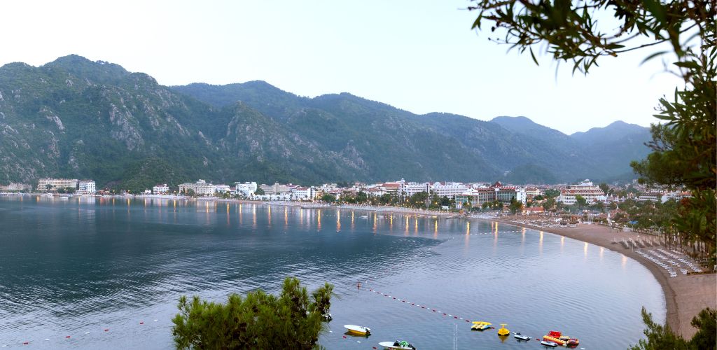 A beach surrounded by green mountains and buildings and a few small boats floating at the near shore. 