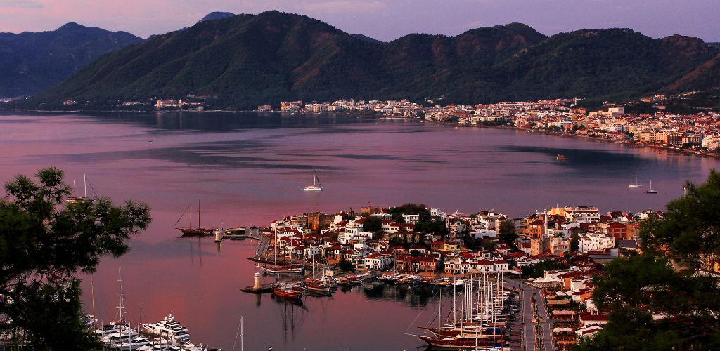 A view of the sea, as well as buildings and mountains. A yacht parked on the beach. 
