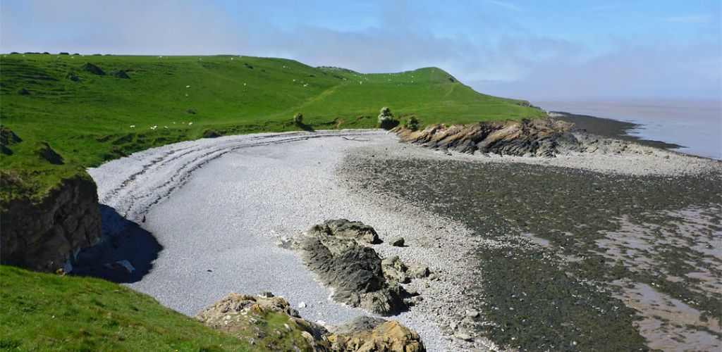 At the clear sky and sea water, there is a green mountain and gray sand. 
