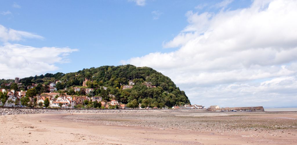 A large green mountain with residences on top. A low tide beach scene with a cloudy sky.  
