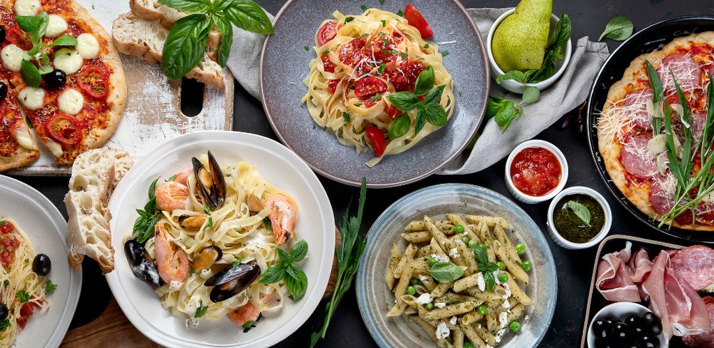 On the table are different kinds of pasta, pizza, sauces, and hams. 