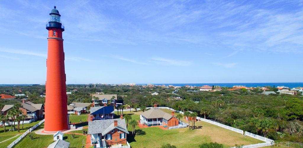 Lighthouse and houses on Daytona Beach, green trees, and a clear sky. 
