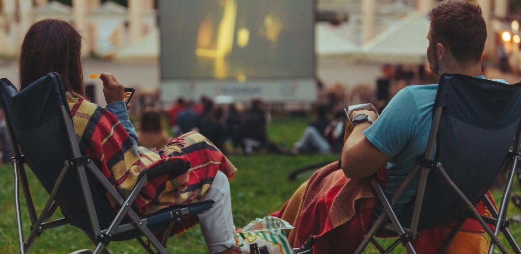 A couple eating chips while watching movies at the open park with another group of people in the background. 