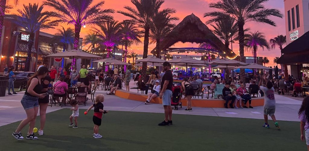 A crowd of people relaxing in a tiny tent and playing with children in the center, surrounded by building stores, restaurants, and trees while the sun sets. 