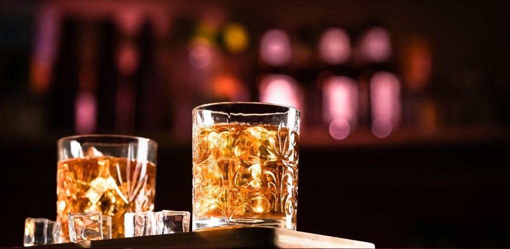 Two glasses of whiskey with an ice cube on the table beside the glass.