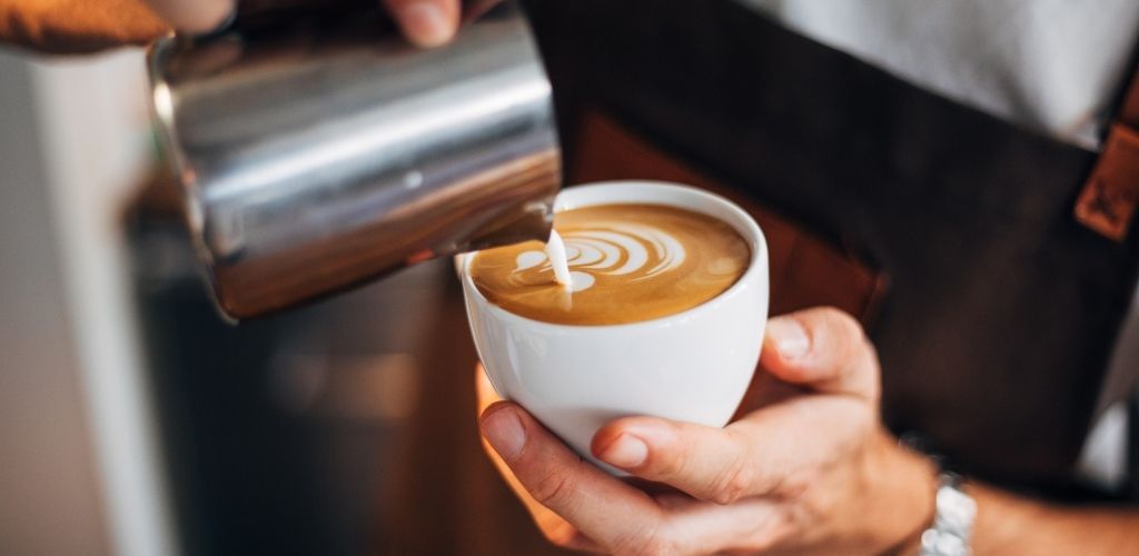 A barista hold a cup of latte and a stainless with milk.