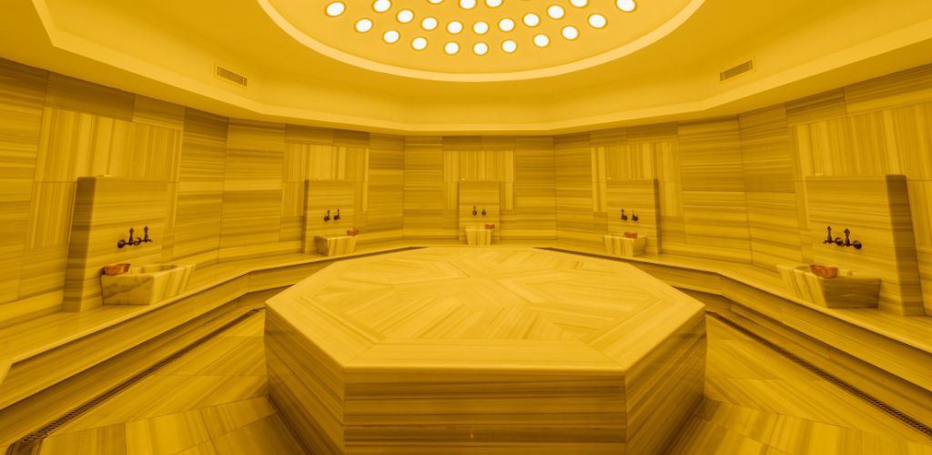 A bath with marble stone shape octagon at the center where you can laydown and surrounded a marble stone sink with yellow dim light.