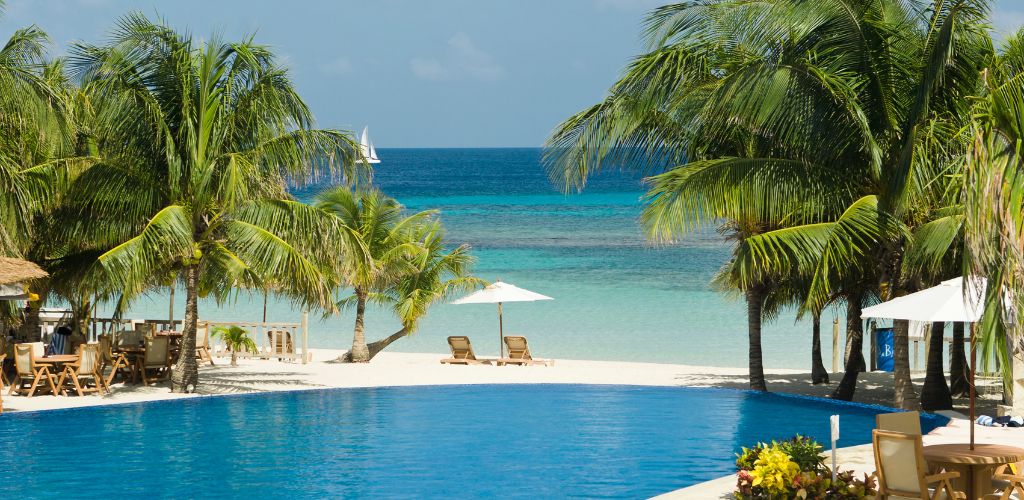 There is a pool surrounded by coconut trees, with tables and seats on the side. There is white sand on the beach and two beach beds in the background.