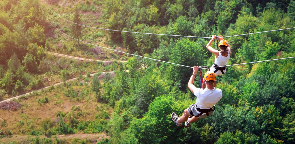 Zipline is an exciting adventure activity. Man and woman ride on the Zipline cable car with helmets and the green trees underneath. 