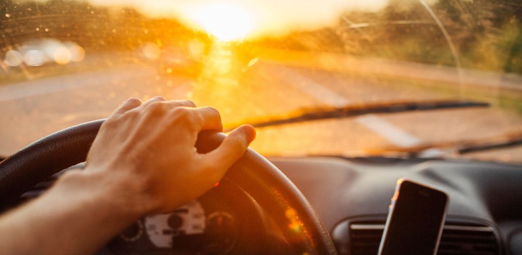 A man's hand holding a car wheel and a cell phone in the front, with a sunset light on the windshield. 