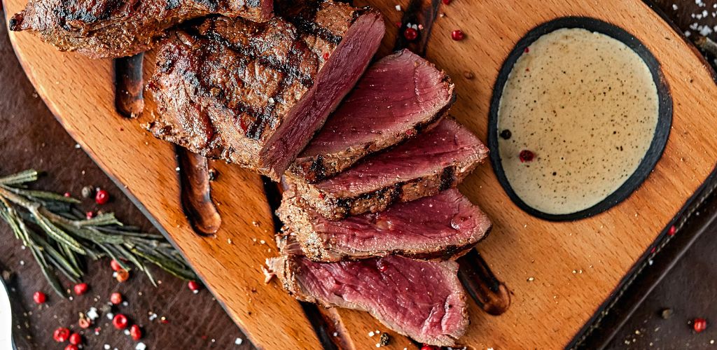 On the wooden plate, there is a cut steak and steak sauce. 