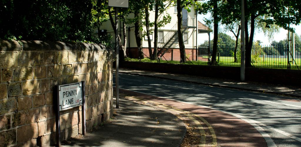 Penny Lane sign in Liverpool road and there is a white house on the background and green field and trees. 