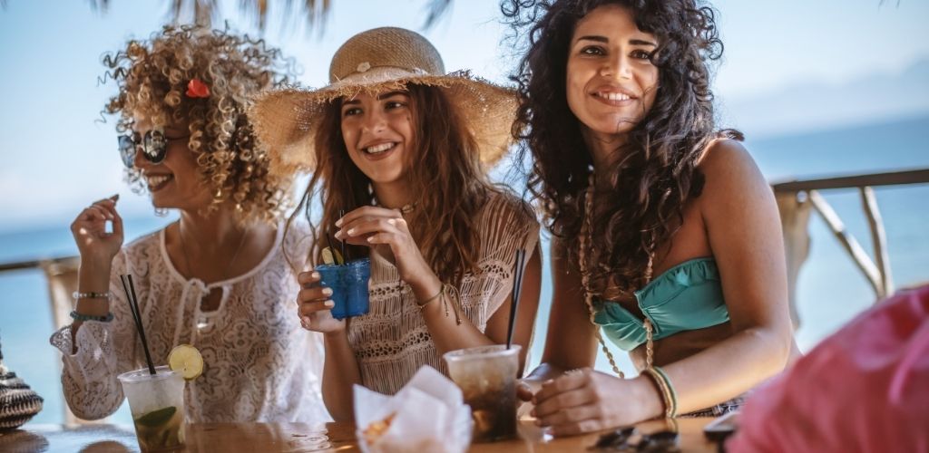 Three women on the bar beach holding a cocktail drink. 