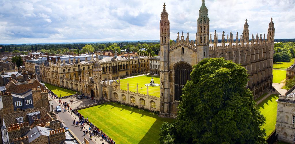 A view of Cambridge University and King's College Chapel, a large green tree, a building structure on the opposite side of Cambridge University, and a crowded people on the street. 
