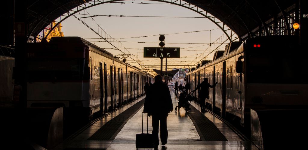 A shallow shot of a man holding his travel luggage at a train station, with two trains in the background. 
