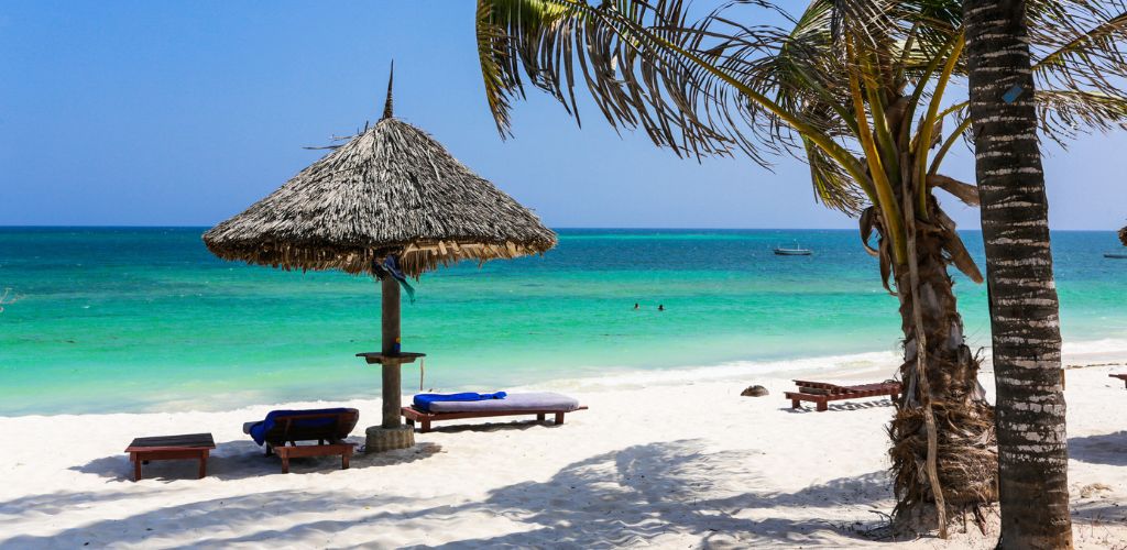 The beach has white sand. There is a modest pavilion on the beach and four beach beds. There are also two coconut trees on the side. 