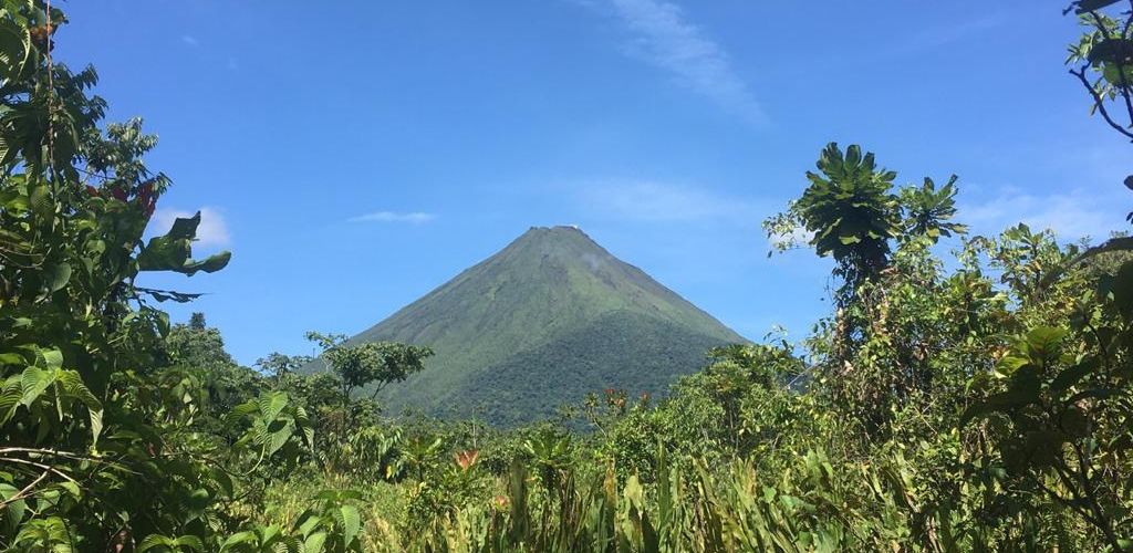 A distant image of a volcano with green plants surrounding it. 