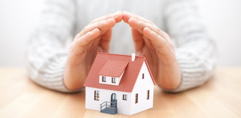 On a wooden table, a model house and a hand are protecting the model house. 