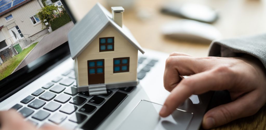 A hand of a man and a small doll house on the laptop and a house on the screen. 