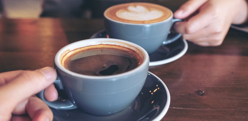 Close-up image of a man and a woman clinking blue coffee mugs on a wooden table in a cafe. 